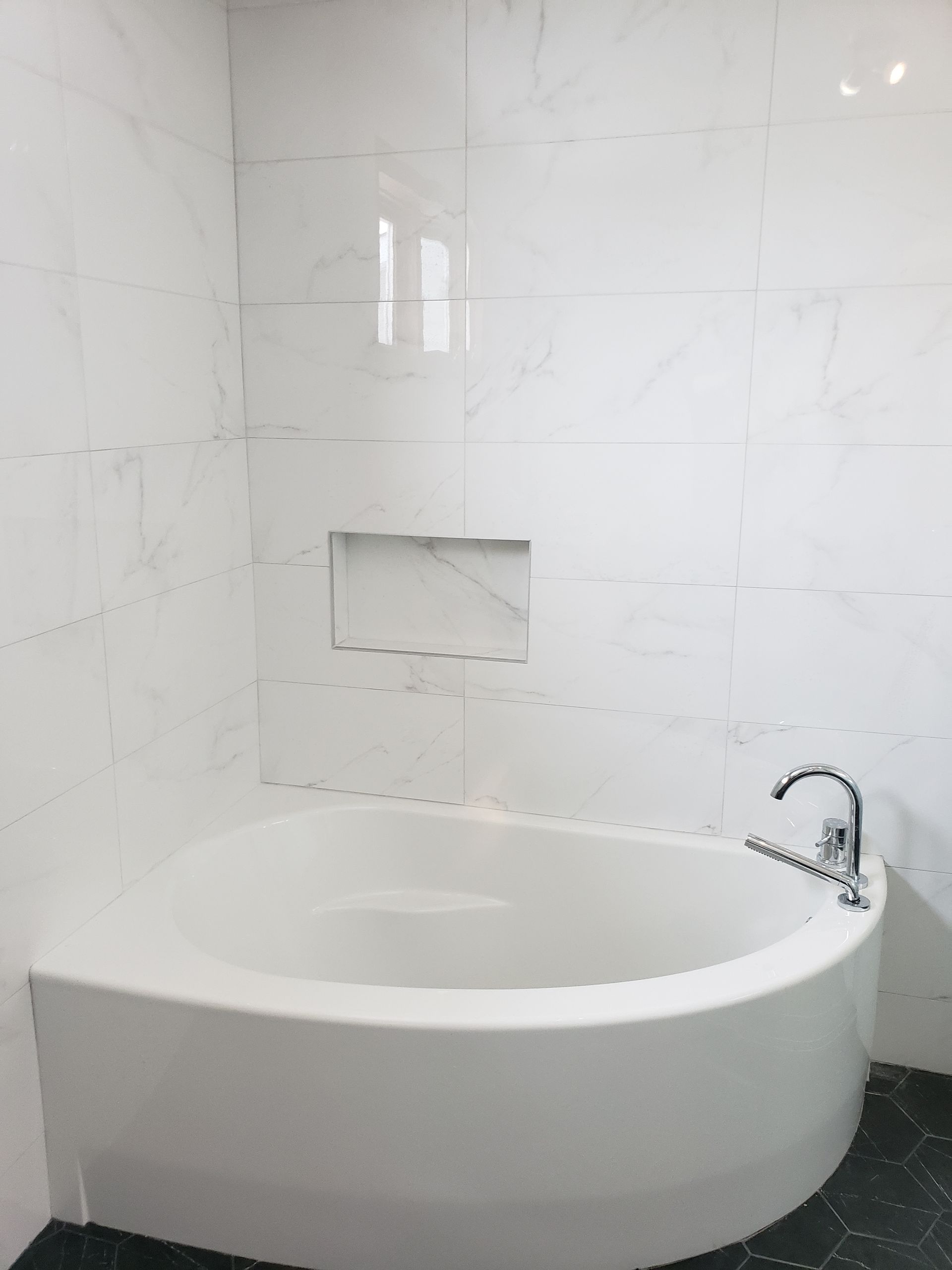 Baignoire d'angle blanche dans une salle de bains carrelée avec étagère intégrée.