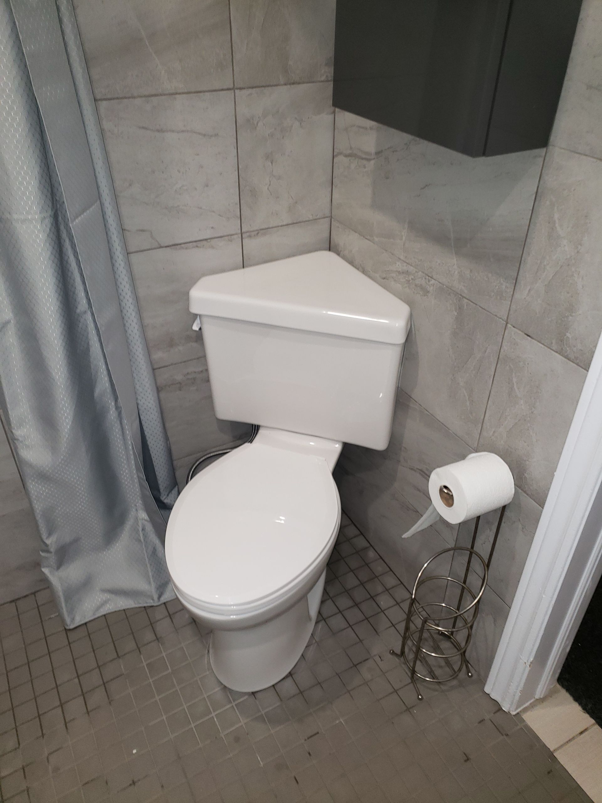 Toilettes d'angle blanches dans une petite salle de bains carrelée de gris avec un porte-papier toilette.