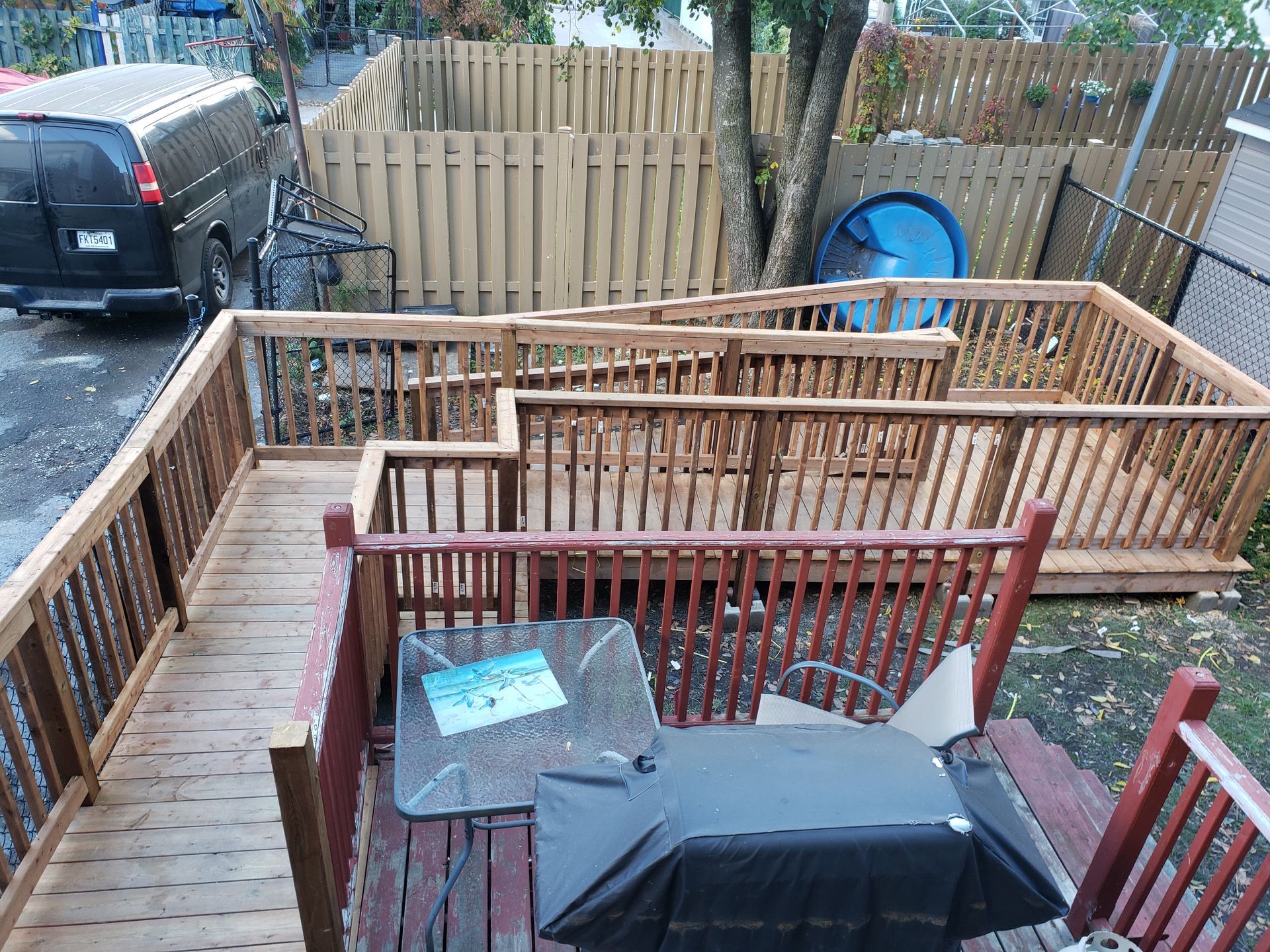 Terrasse en bois à plusieurs niveaux, barbecue, table et haute clôture en bois. Fourgonnette garée à gauche.