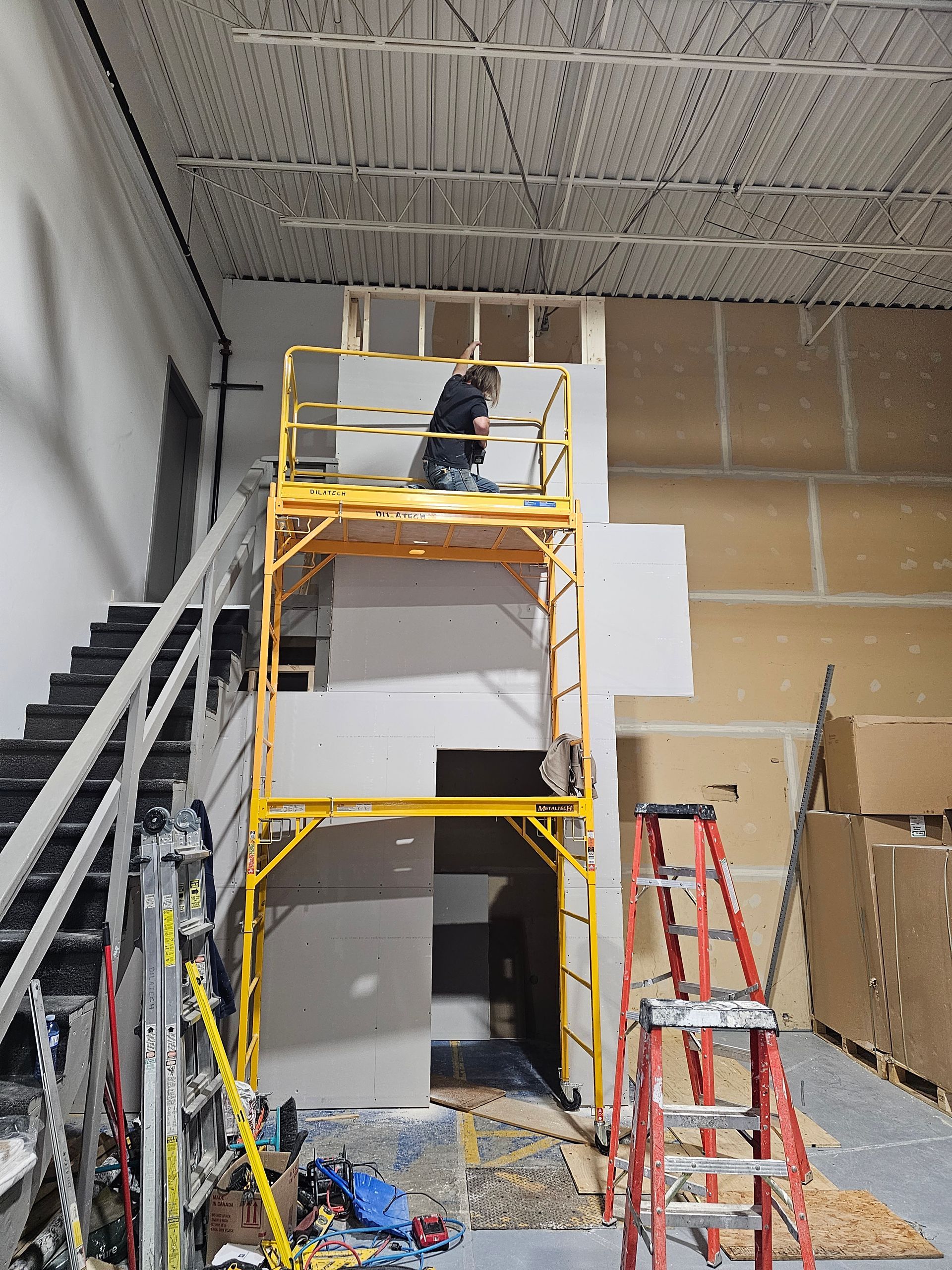 Une personne sur un échafaudage jaune construit un mur dans un bâtiment avec des escaliers et une autre échelle.