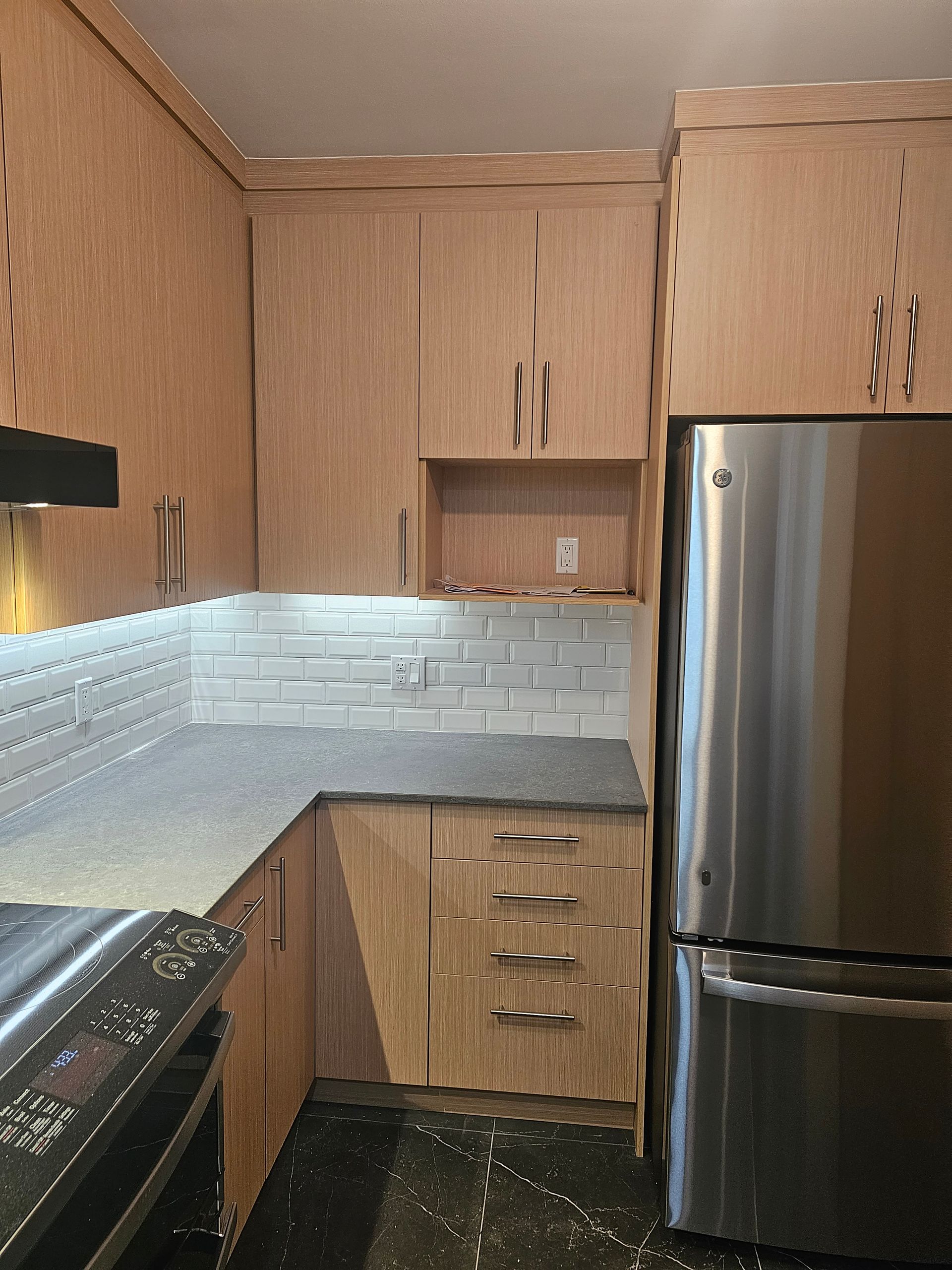 Cuisine avec armoires en bois clair, comptoirs gris, crédence en carrelage blanc et réfrigérateur en acier inoxydable.