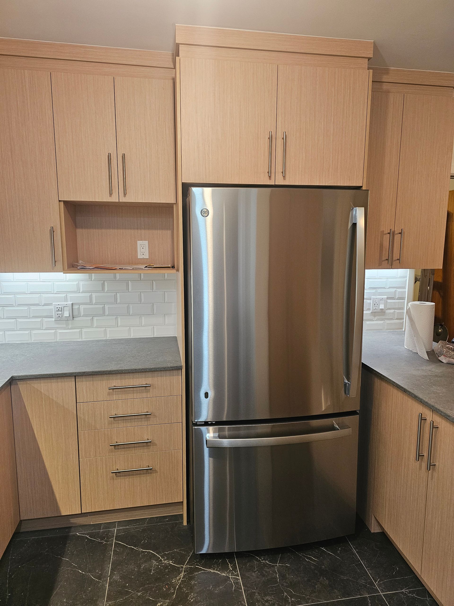 Cuisine avec placards en bois clair, réfrigérateur en acier inoxydable et plans de travail foncés.