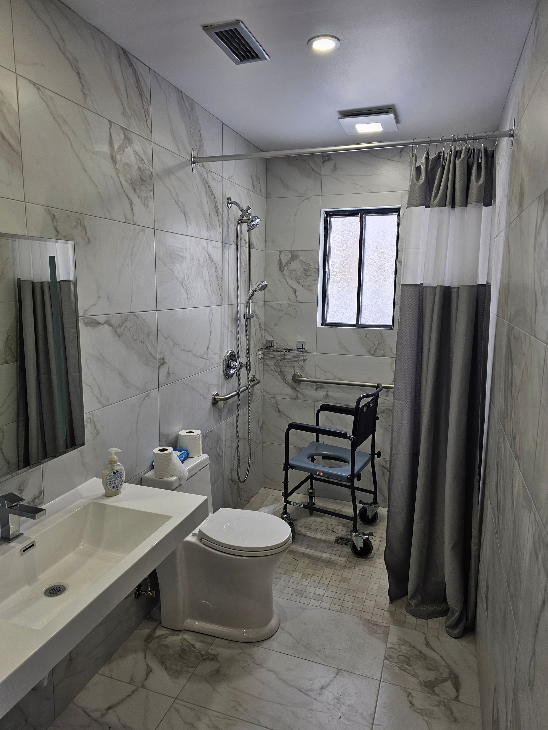 Salle de bains accessible avec douche, lavabo, toilettes et emplacement pour fauteuil roulant. Murs et sol en marbre, rideau de douche gris.