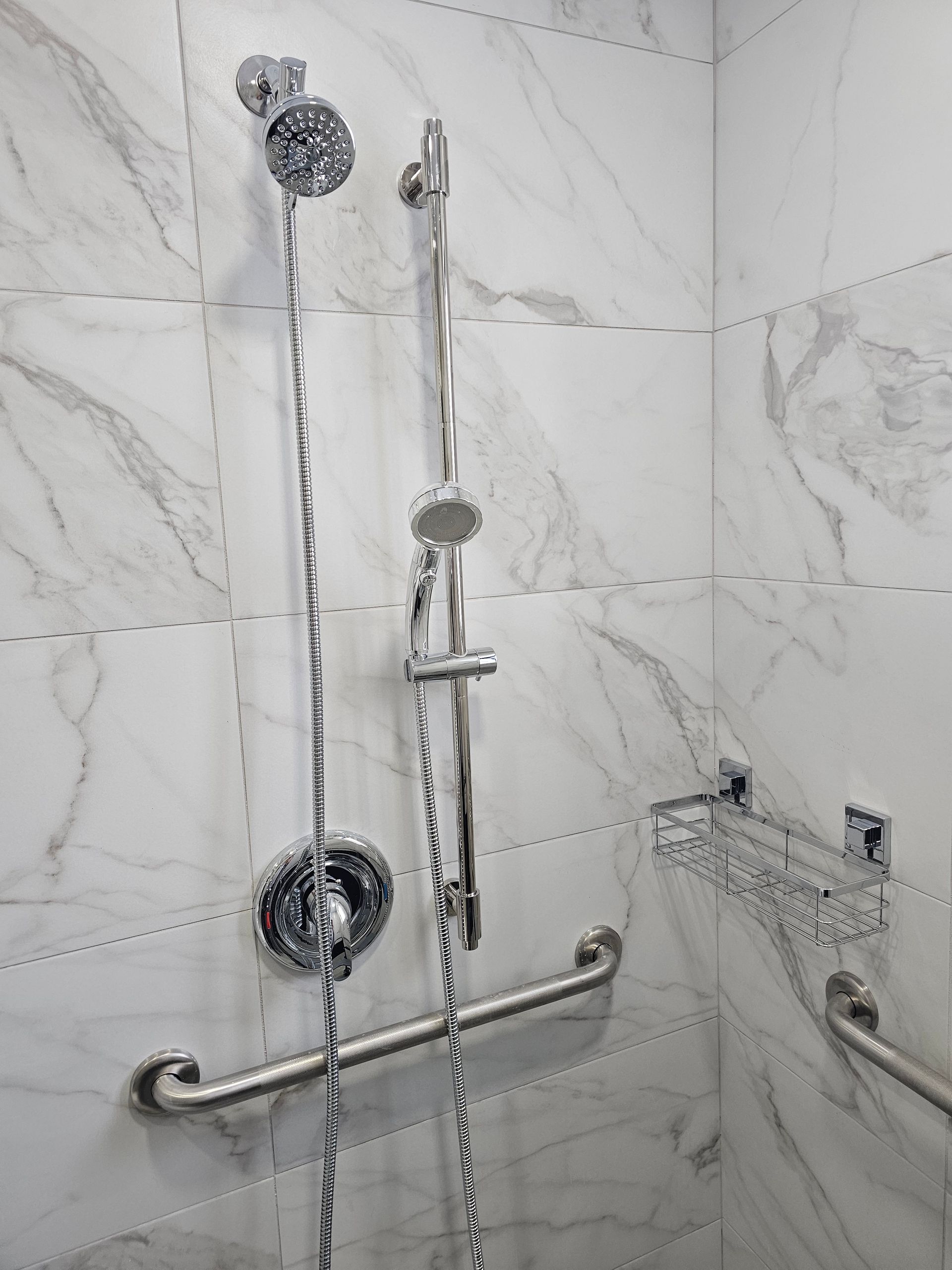 Une douche carrelée de blanc avec pommeau de douche, douchette, barres d'appui et porte-savon.