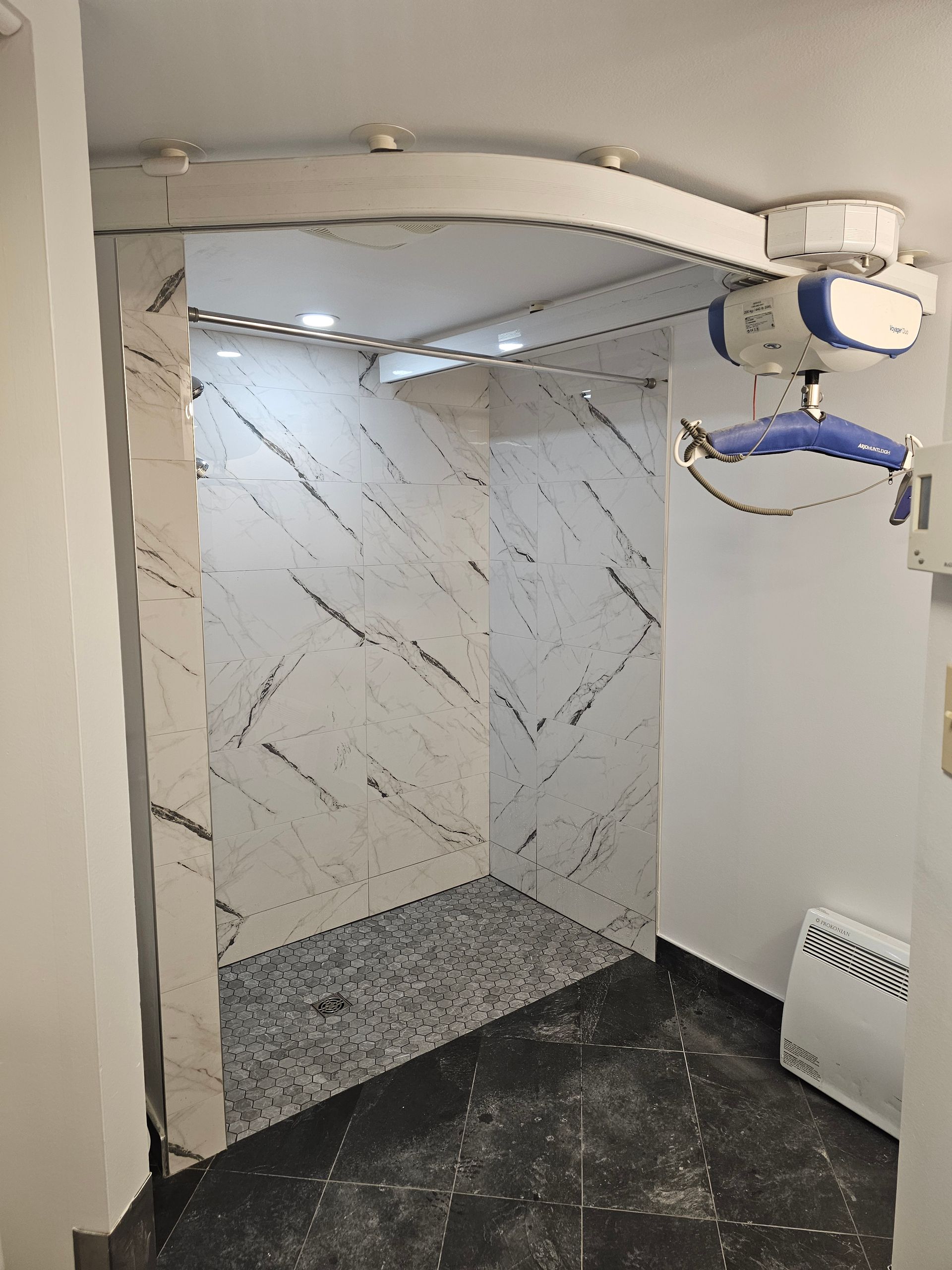 Petite cabine de douche aux murs à motif marbre et au sol foncé. Les équipements fixés au plafond sont visibles.