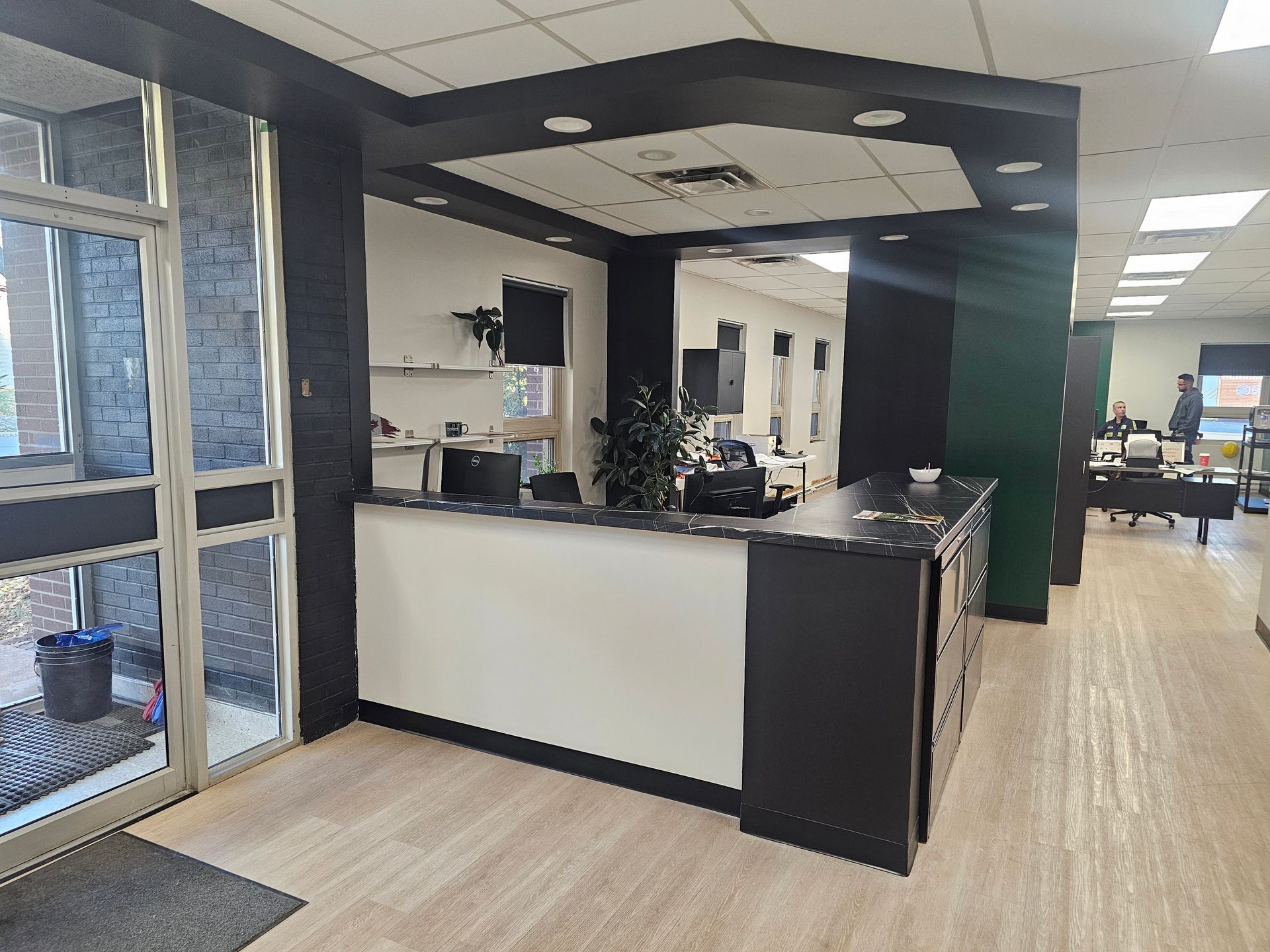 Réception dans un bureau. Comptoir noir et blanc avec une petite plante. Porte ouverte à gauche, couloir en arrière-plan.