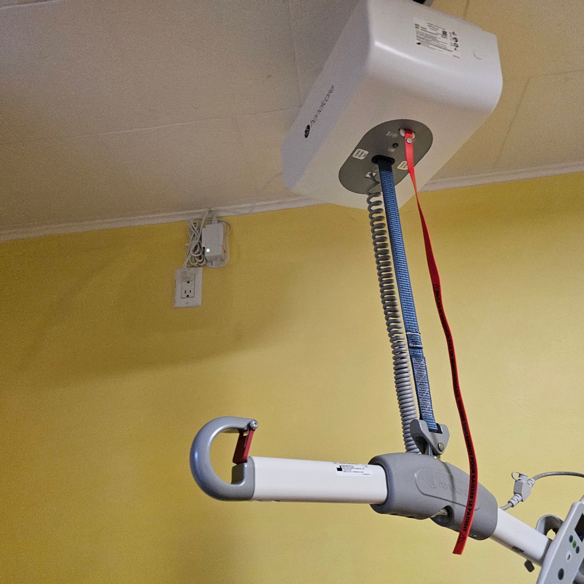 Lève-personne fixé au plafond, gris et blanc, avec sangles bleues et rouges, installé dans une chambre.