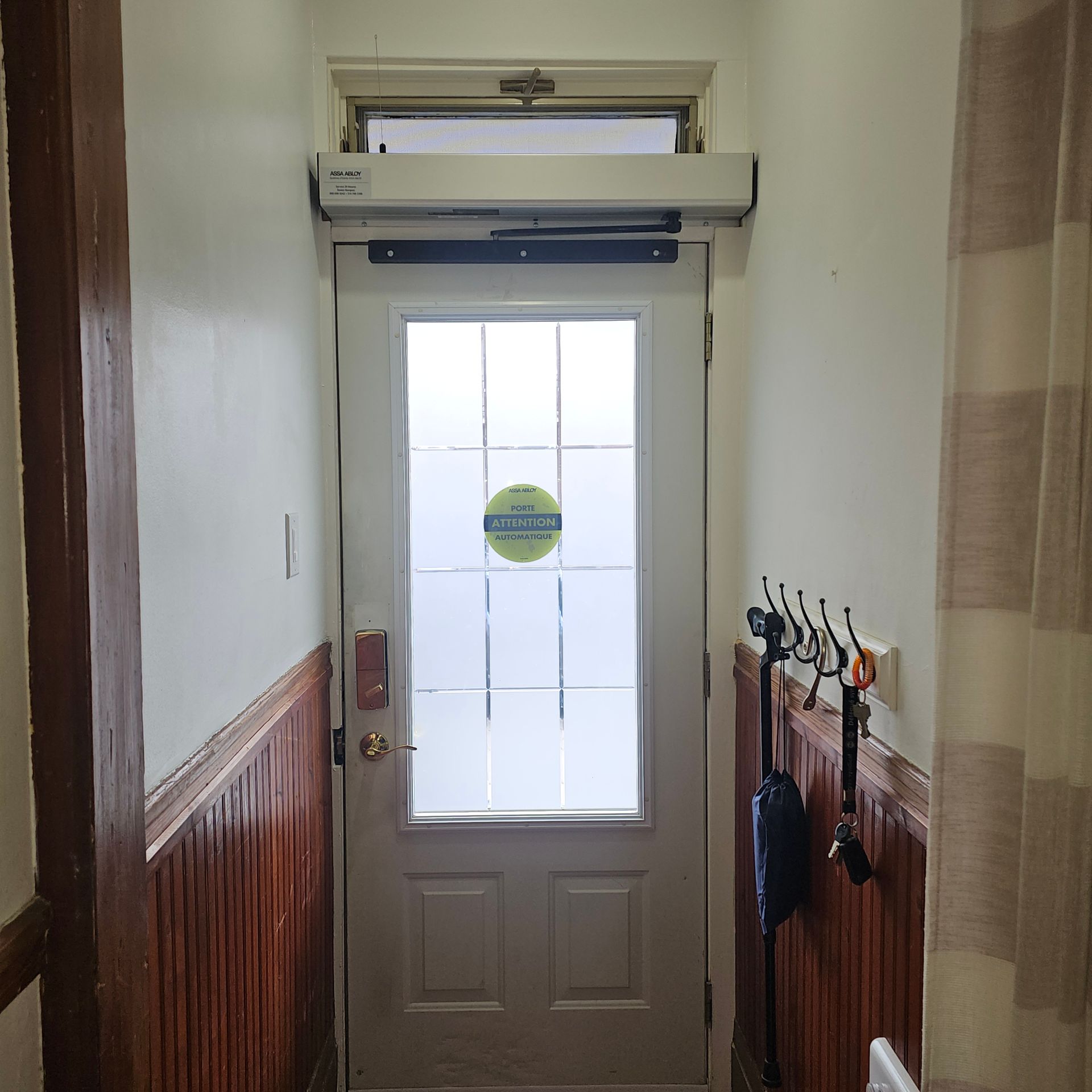 Porte blanche avec panneau en verre dépoli, ouvre-porte automatique, couloir avec lambris, porte-manteau et rideau.
