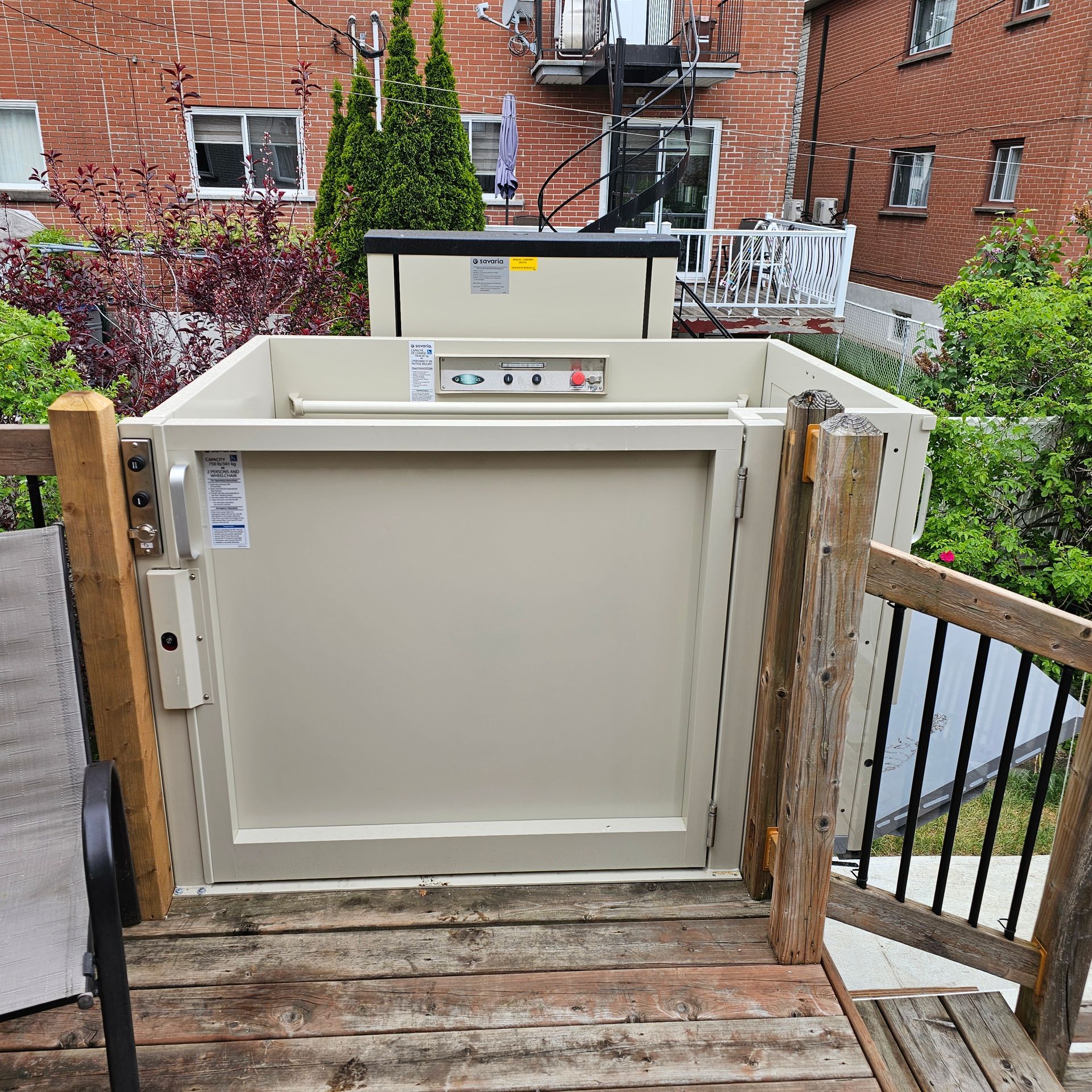 Ascenseur vertical beige sur une plateforme en bois, devant un bâtiment en briques rouges.
