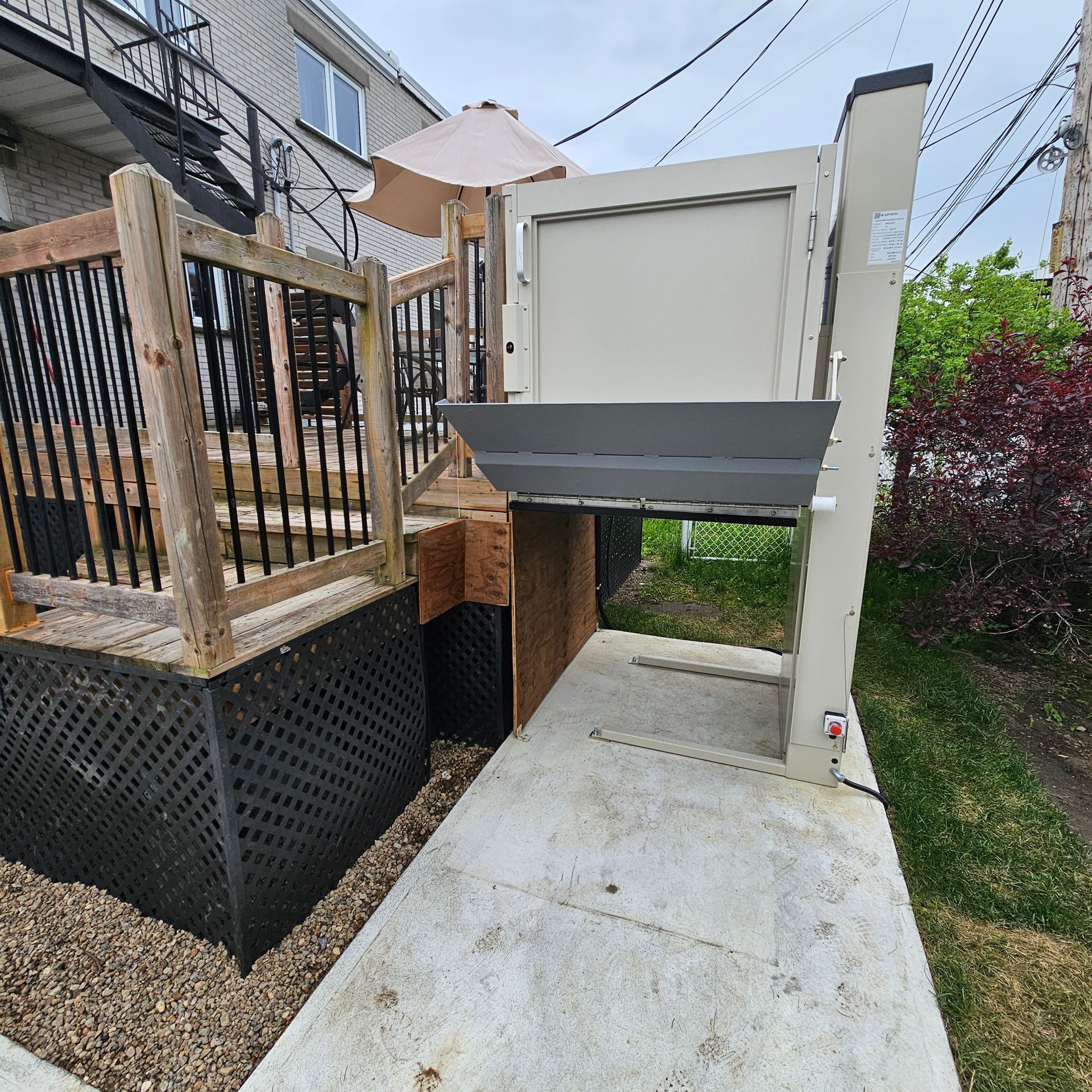 Vue extérieure d'un élévateur pour fauteuil roulant beige, d'une rampe en béton et d'une terrasse en bois à côté d'un bâtiment.