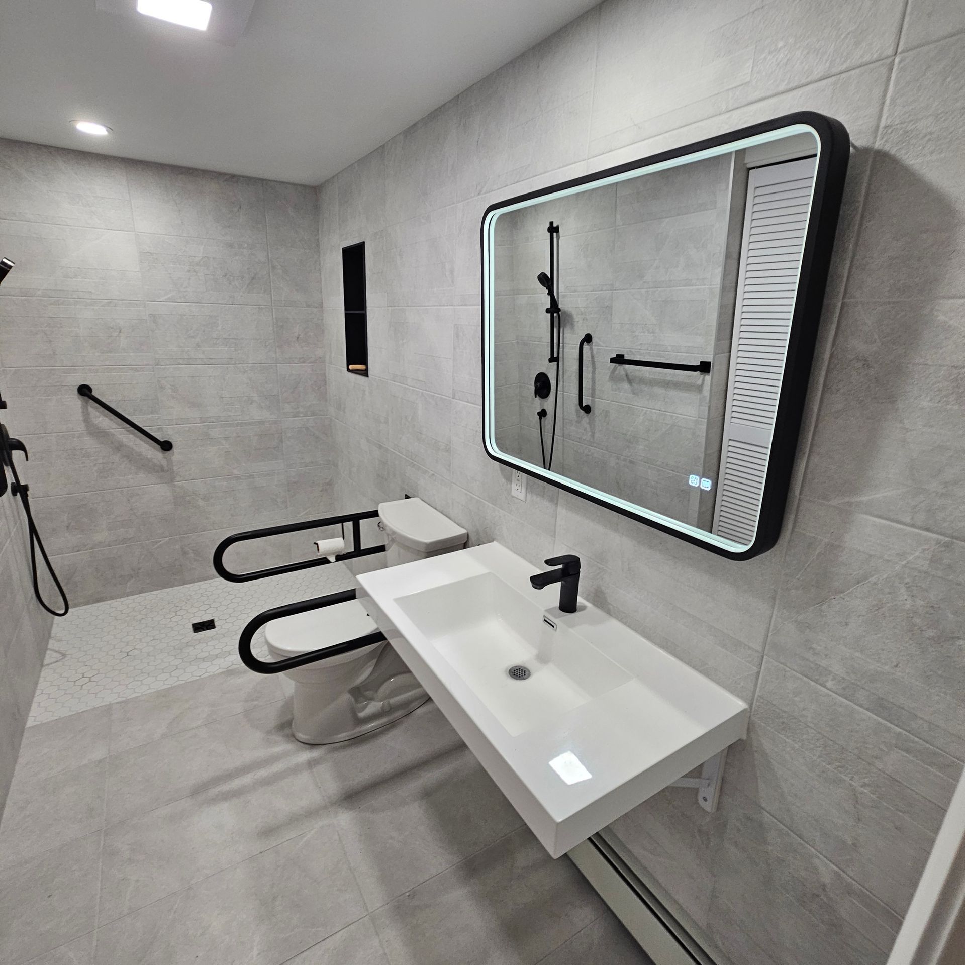 Salle de bain accessible : carrelage gris, lavabo blanc, robinetterie noire, barres d’appui, toilettes avec barres de soutien et grand miroir.