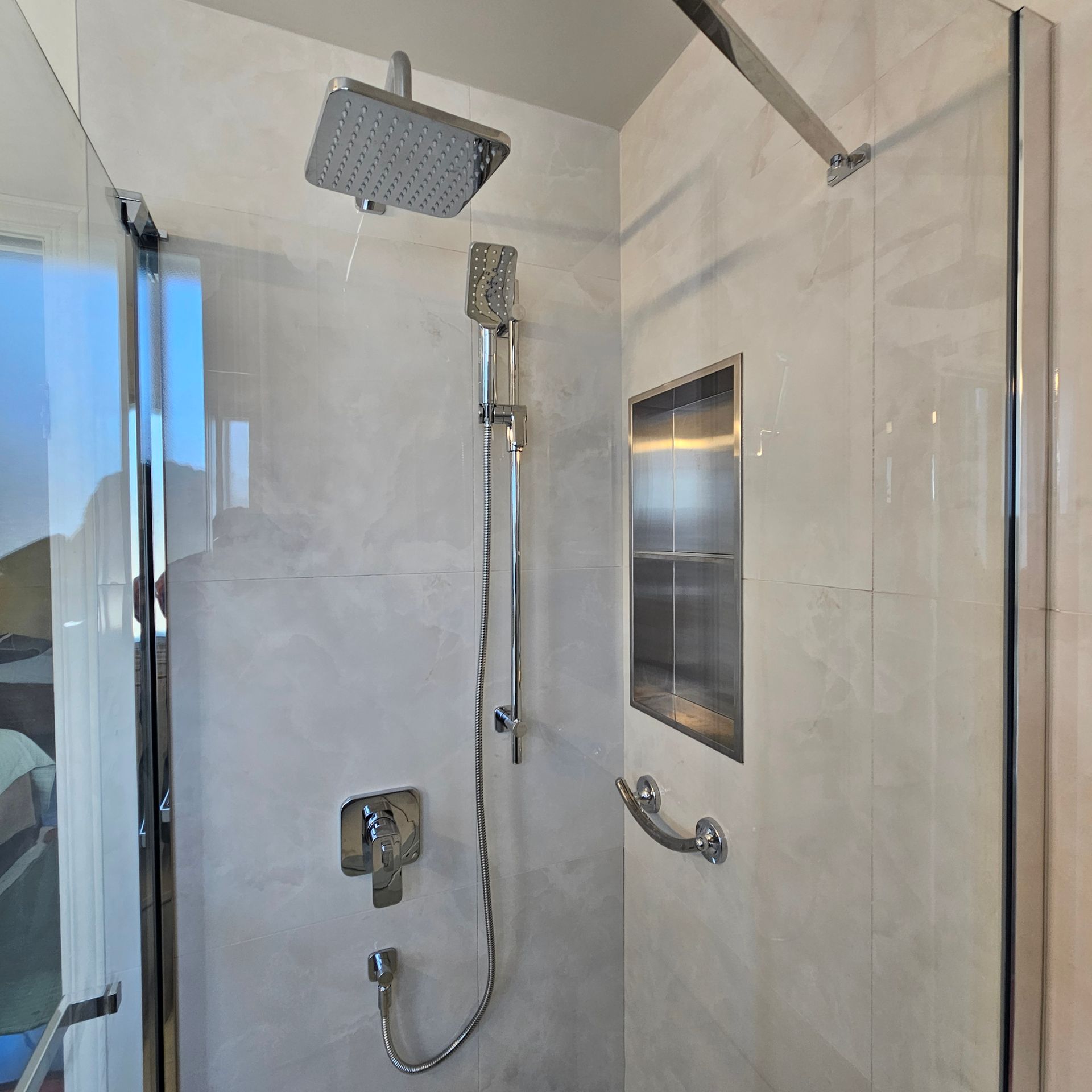 Douche avec pommeau effet pluie, douchette et étagère encastrée dans une cabine carrelée de blanc avec porte en verre.