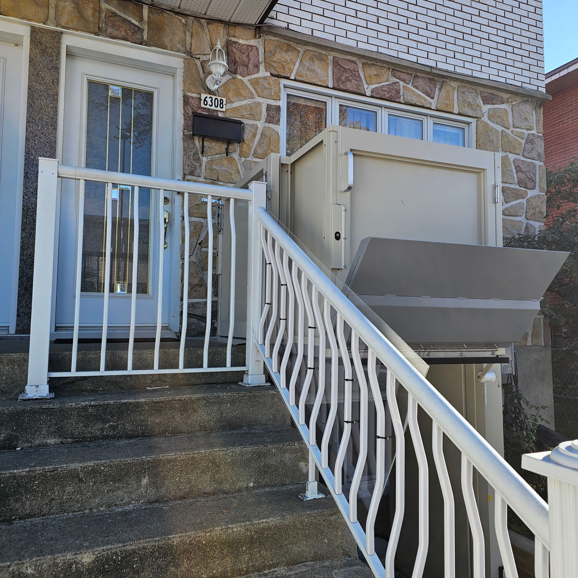 Un immeuble avec un monte-escalier et une rampe blanche sur des marches en béton. Façade en pierre avec porte blanche.