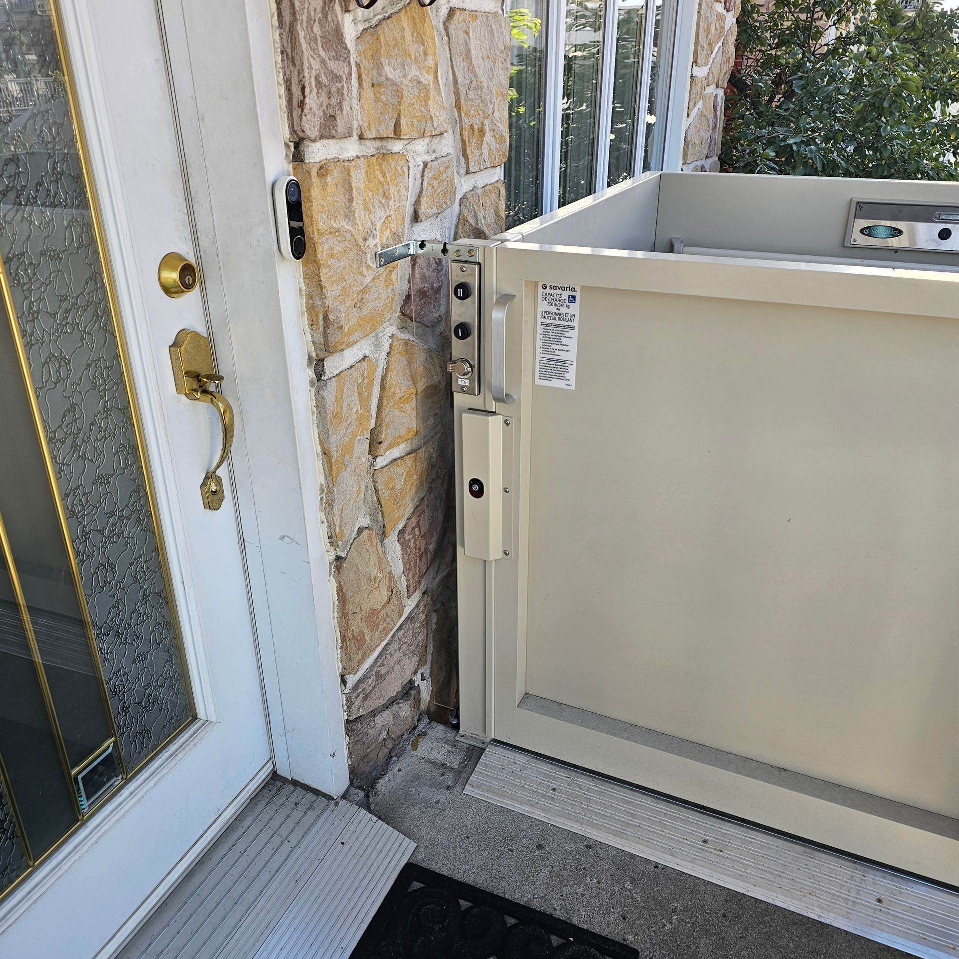 Un ascenseur pour fauteuil roulant beige se trouve à côté de l'entrée d'une maison ; la porte est à gauche, un mur en pierre se trouve derrière l'ascenseur.