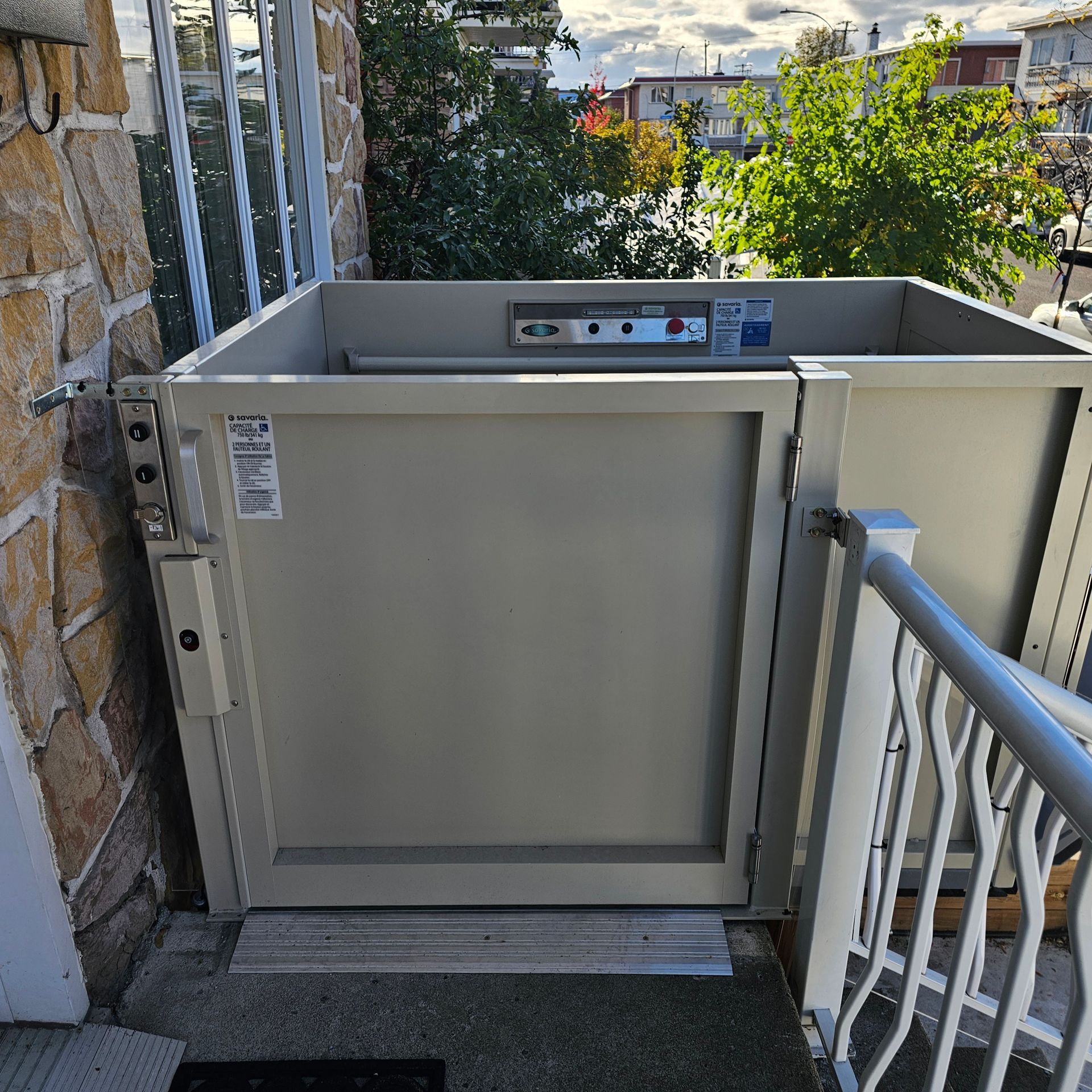 Un ascenseur beige est installé sur le porche d'un bâtiment en pierre. Un portail et une rambarde métalliques se trouvent à proximité. Des arbres sont visibles en arrière-plan.
