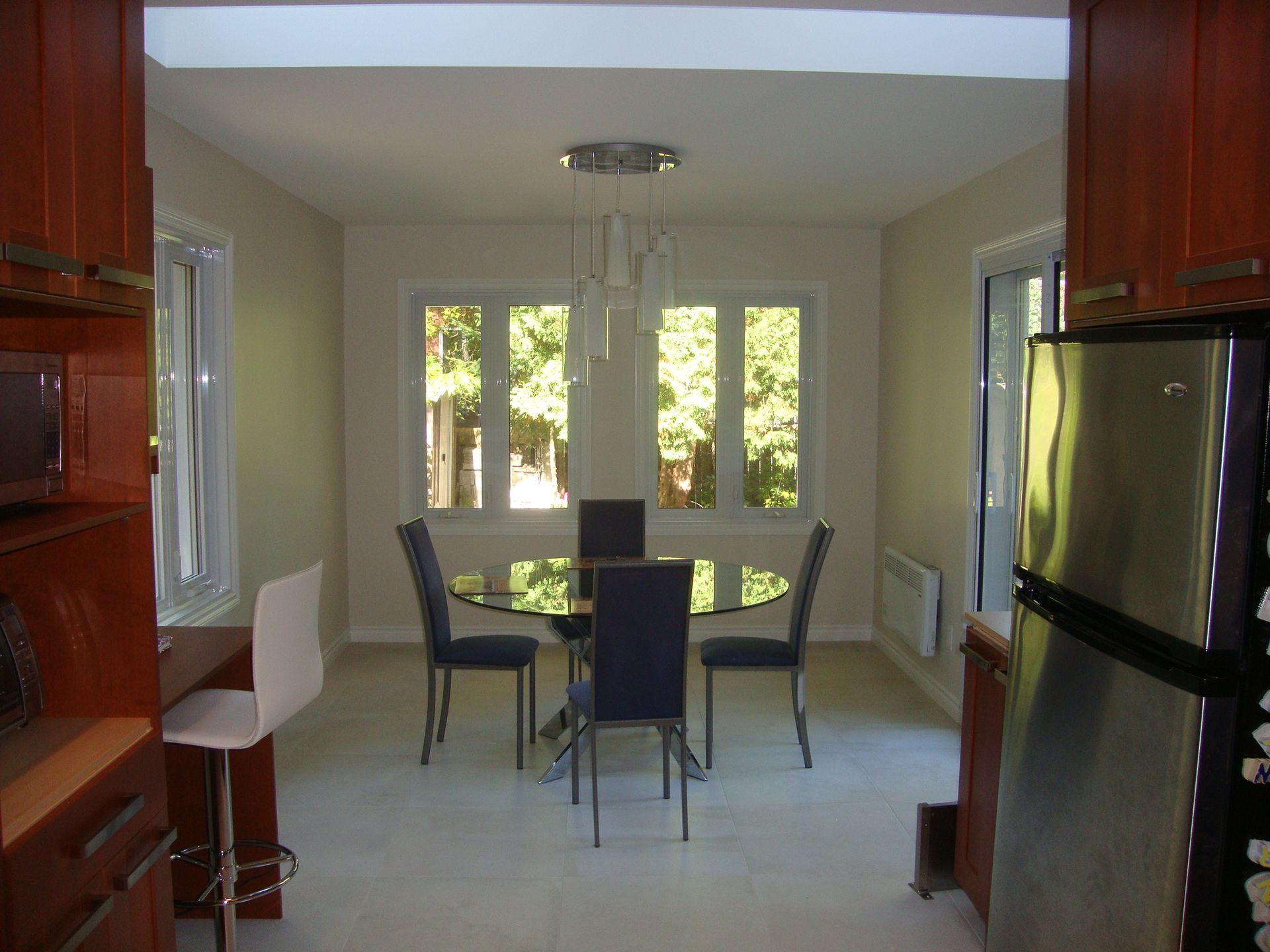 Salle à manger avec table en verre, quatre chaises bleues, deux fenêtres et luminaire moderne.