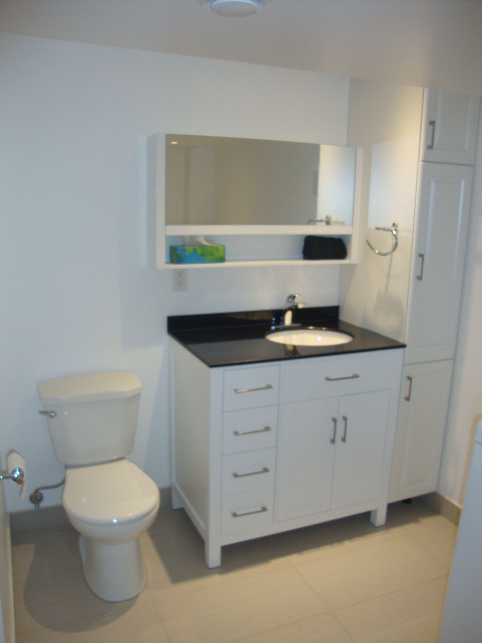 Salle de bains avec toilettes, meuble-lavabo et armoire blancs sur des murs blancs. Plan de travail et miroir noirs.