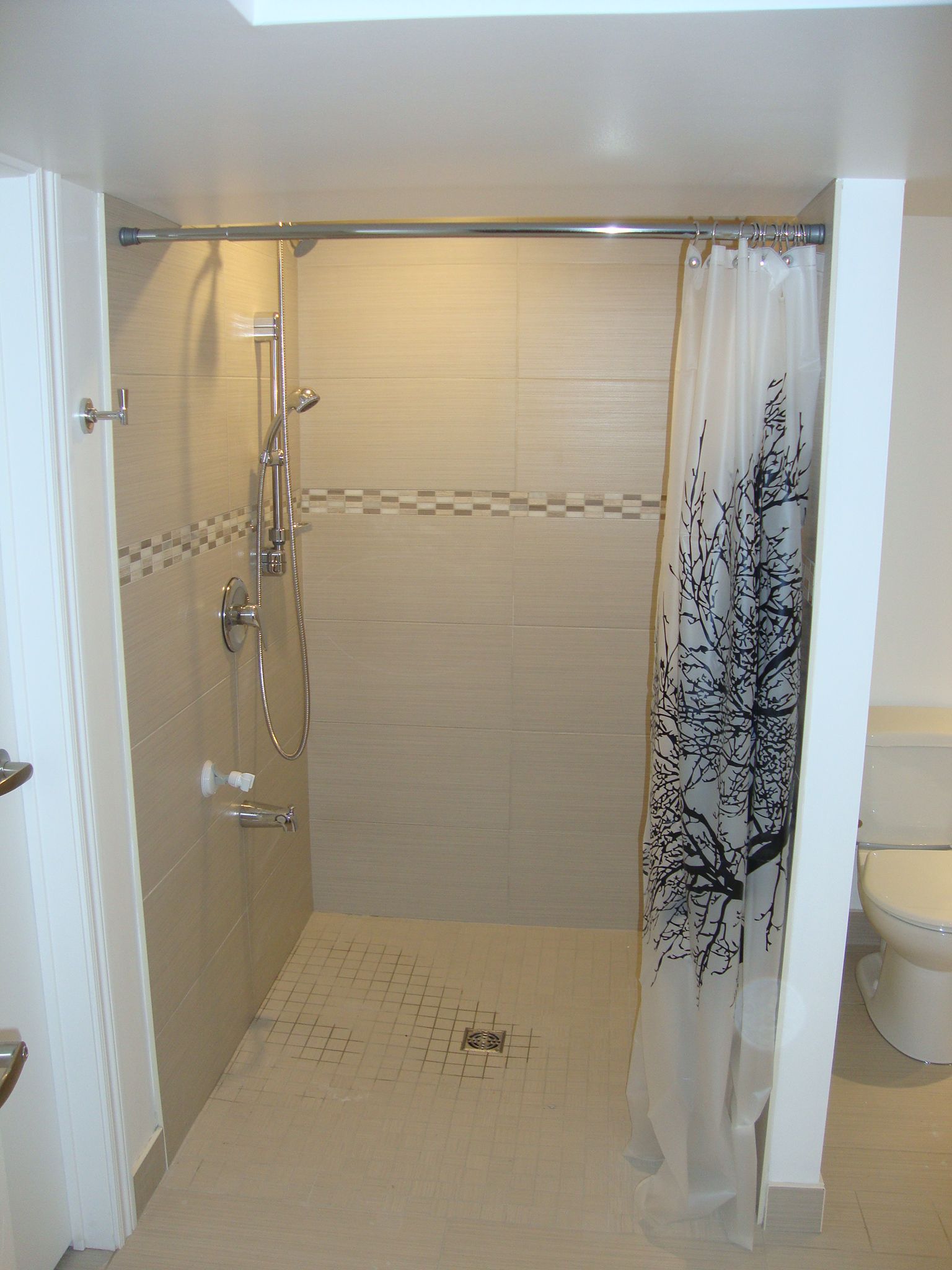 Douche avec carrelage blanc et gris, pommeau de douche argenté et rideau de douche à motifs noirs et blancs.