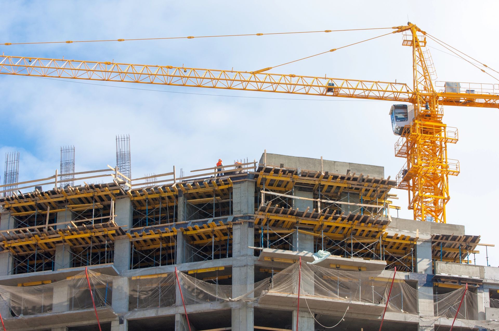 Cantiere con gru gialla che solleva materiali verso un edificio in cemento in costruzione sotto un cielo parzialmente nuvoloso.