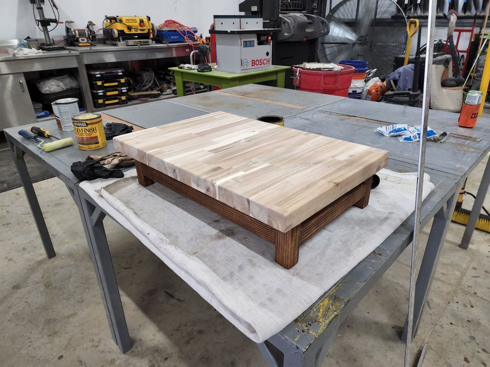 A finished wooden step stool rests on a workbench. The stool has a light-colored top and a darker stained base.