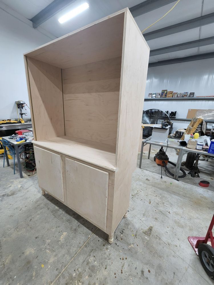 Wooden cabinet under construction in a workshop, with a tall open upper section, a lower cabinet with doors, and small bun feet.