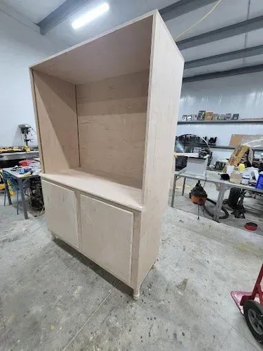 A large, unfinished wooden cabinet stands in a workshop. It has a closed lower section with doors and an open upper section.