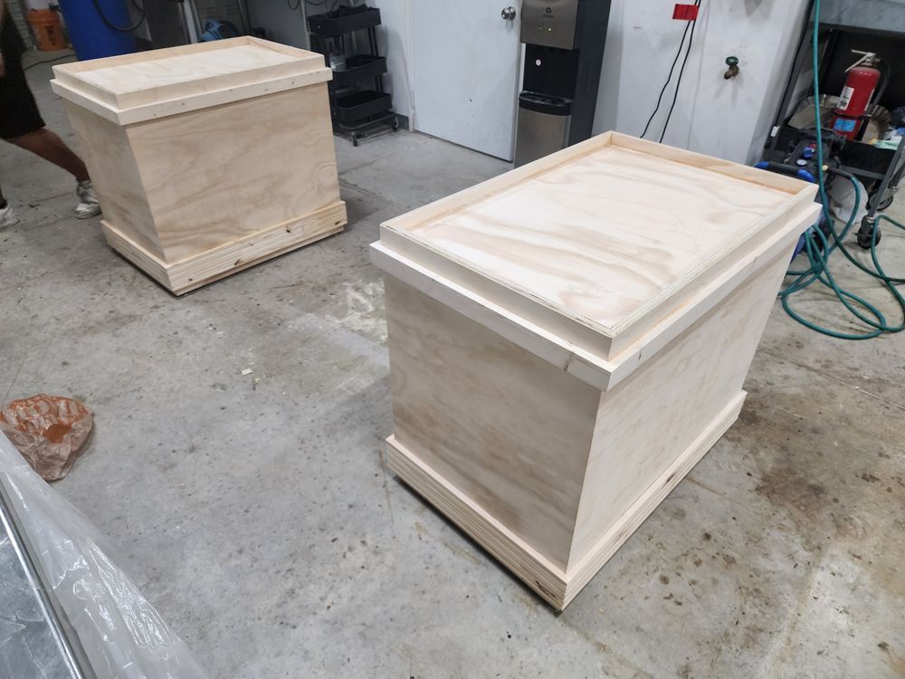 Two unfinished wooden boxes in a workshop setting. They have decorative tops and bases, and sit on a concrete floor.