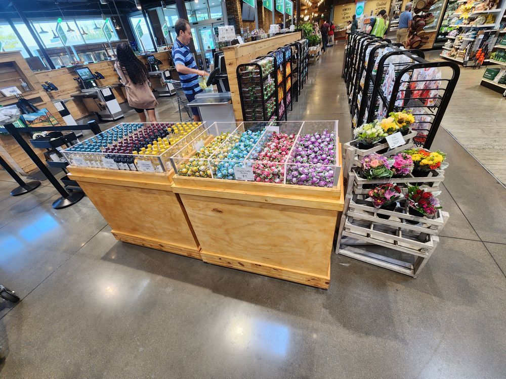 A grocery store display with bins of colorful candy and a flower rack. Shoppers browse the products in a bright, well-lit store.