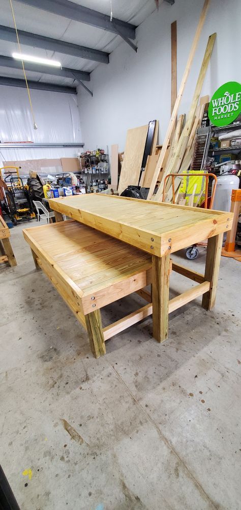 Two wooden workbenches, one above the other, in a workshop. The larger table sits on legs made of posts. The setting has a concrete floor and a cluttered background.