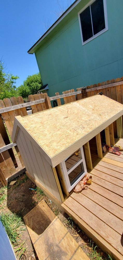 A dog house being constructed on a wooden deck in a backyard, under a blue sky. The house is made of light-colored wood.