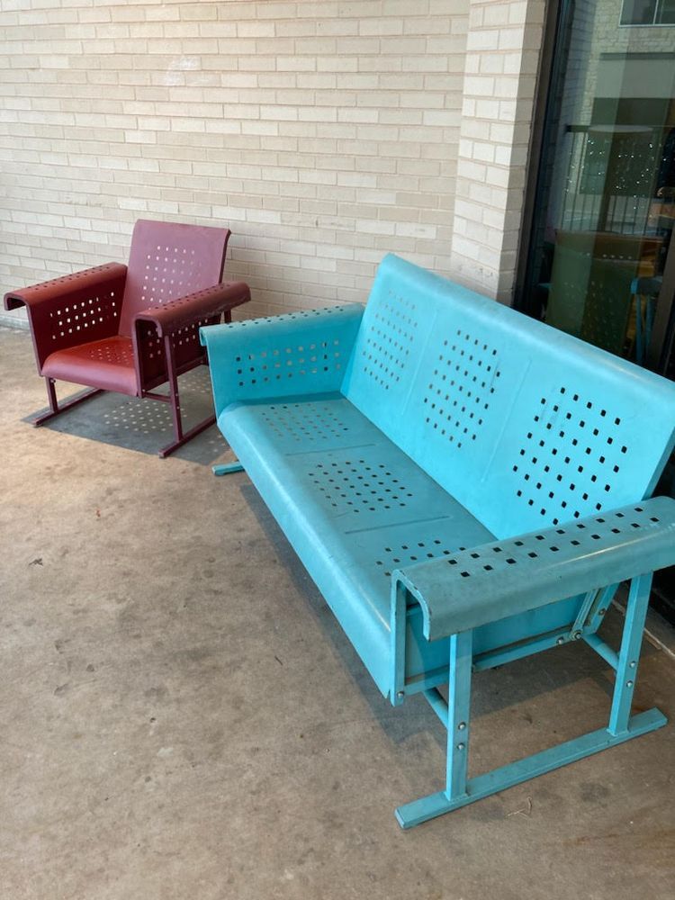 A red chair and a turquoise bench with perforated seats sit on a concrete surface, against a brick wall.