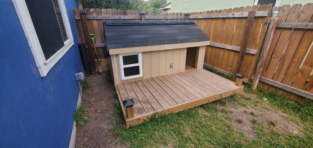 A dog house with a deck in a backyard, next to a blue wall and a wooden fence. The dog house is light brown with a black roof.