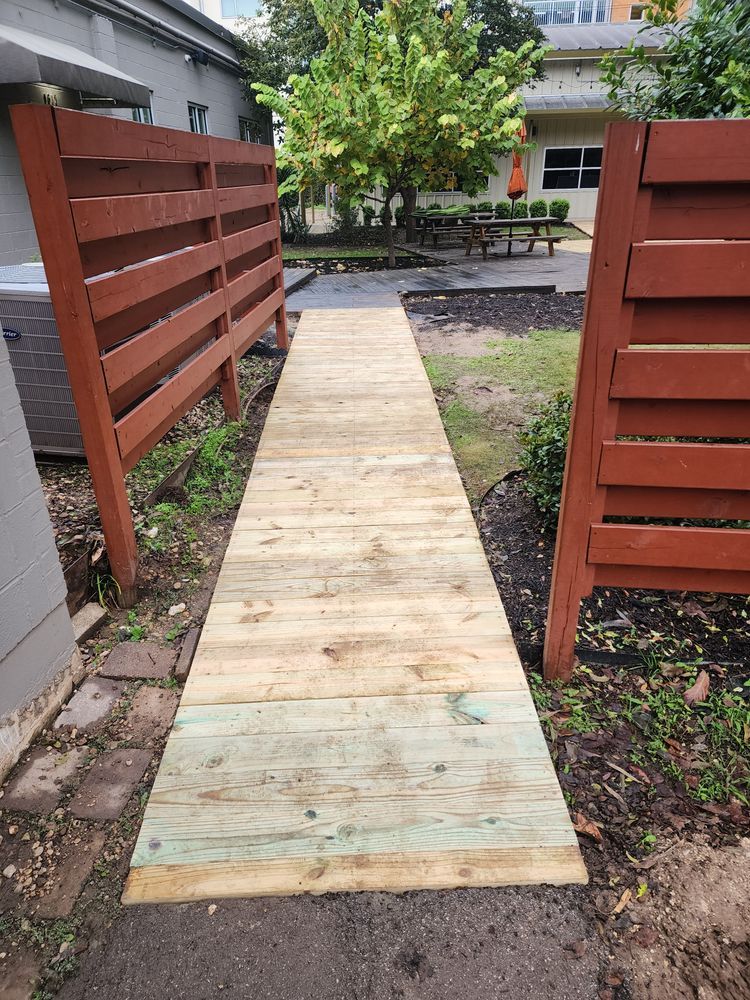 Wooden walkway between two red-brown fences, leading to a patio with tables and a tree. Green and white wood colors.