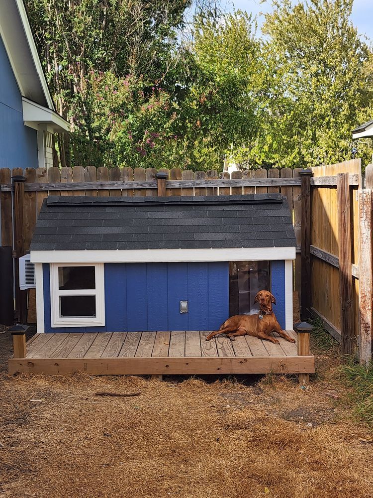 A brown dog relaxes on a wooden deck in front of a blue dog house with a black roof, fenced yard.
