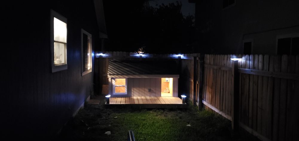 A backyard at night with a lit dog house and string lights along the fences. Two windows of a building are illuminated.