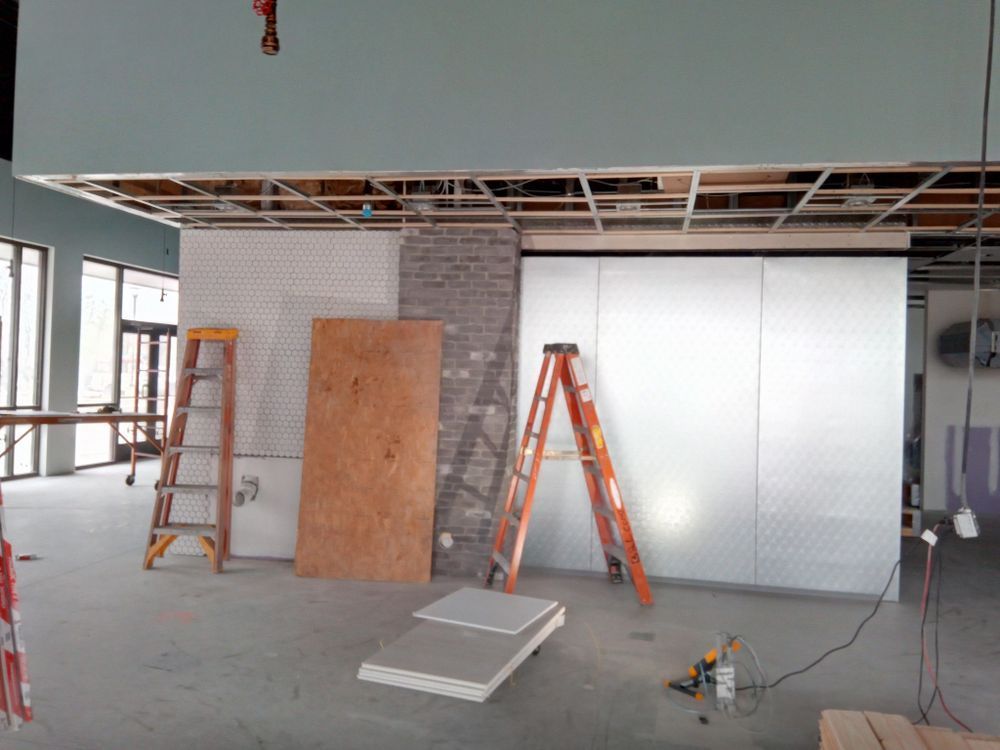 Interior construction site with two ladders, unfinished walls of white tile, brick, and metal panels. The ceiling is partially exposed.