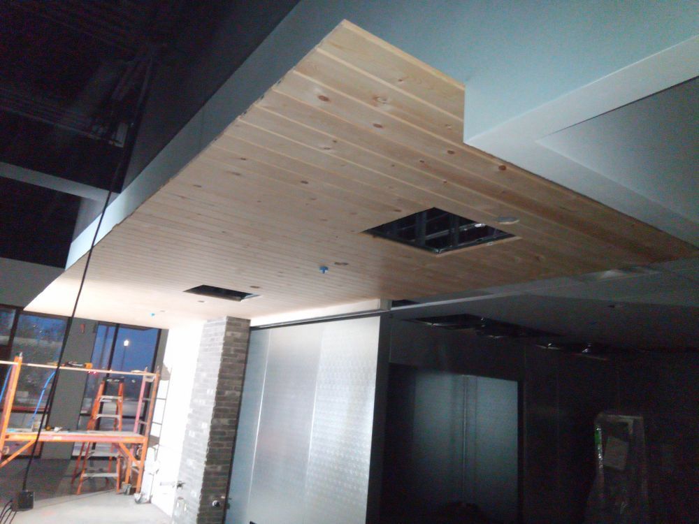 Wooden ceiling with a square cutout, next to a section of light blue ceiling and dark walls; construction in progress.