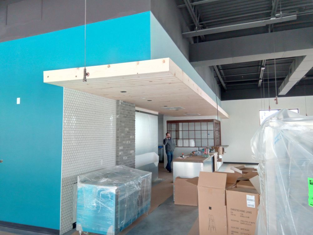 An unfinished commercial space with a long, light-colored counter suspended from the ceiling. A person stands near a stainless steel counter in the background.