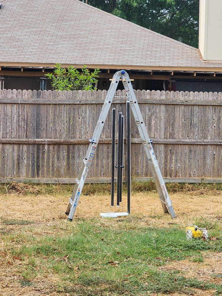 A-frame ladder supporting wind chimes in a grassy backyard setting, with a wooden fence and house roof visible in the background.