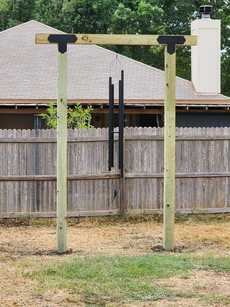 A wooden wind chime stand with black chimes hanging in front of a wooden fence and a house with a chimney.