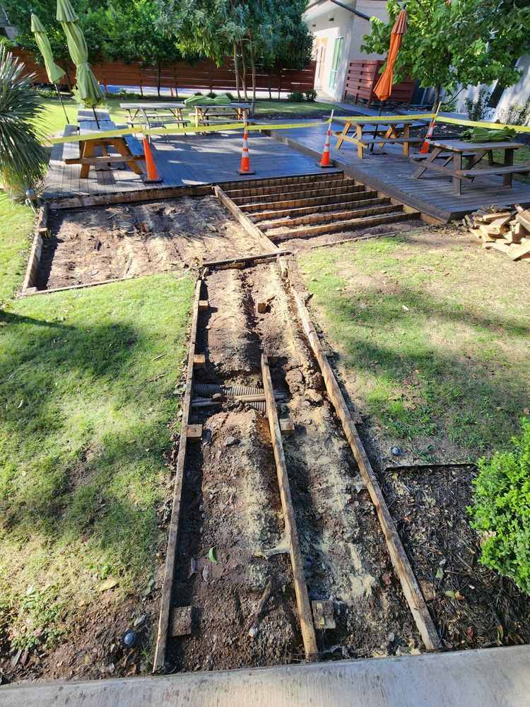 Wooden deck under construction in a grassy outdoor area. The deck boards are removed, revealing the wooden frame. Construction cones and equipment are present.