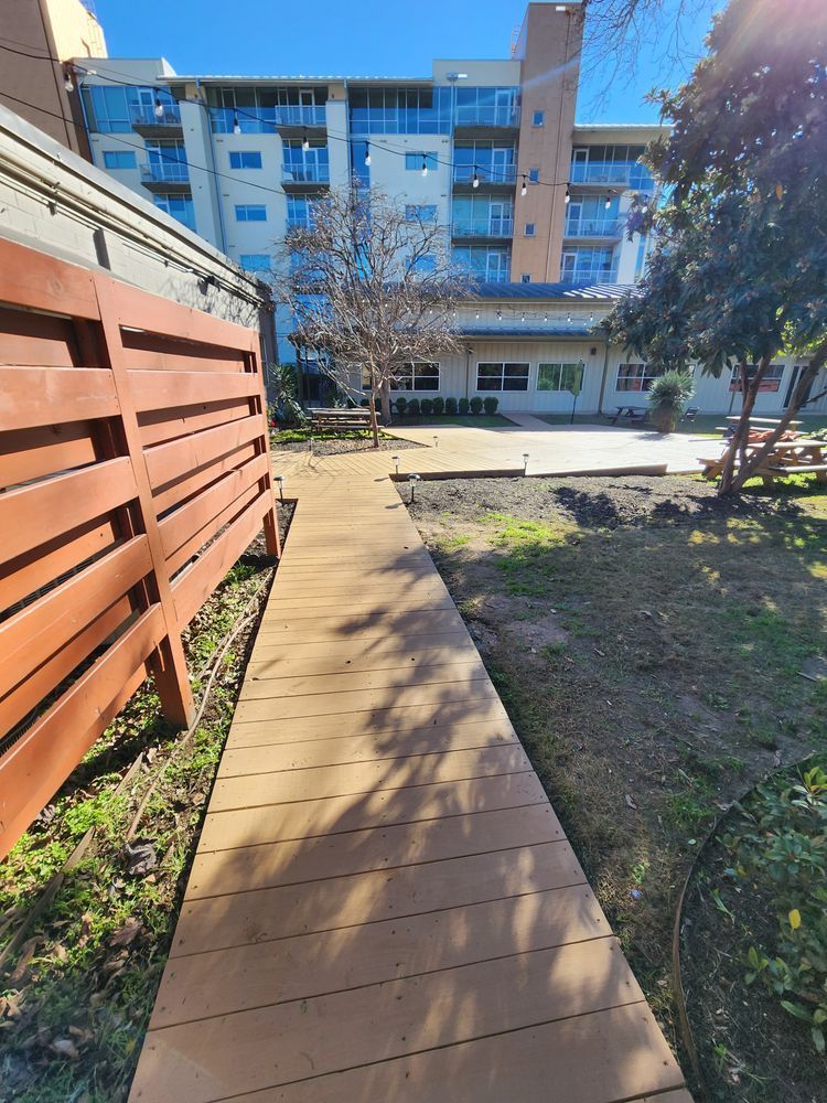 A narrow wooden walkway leads through a yard towards a building in the background. A wooden fence is to the left, and trees are to the right.