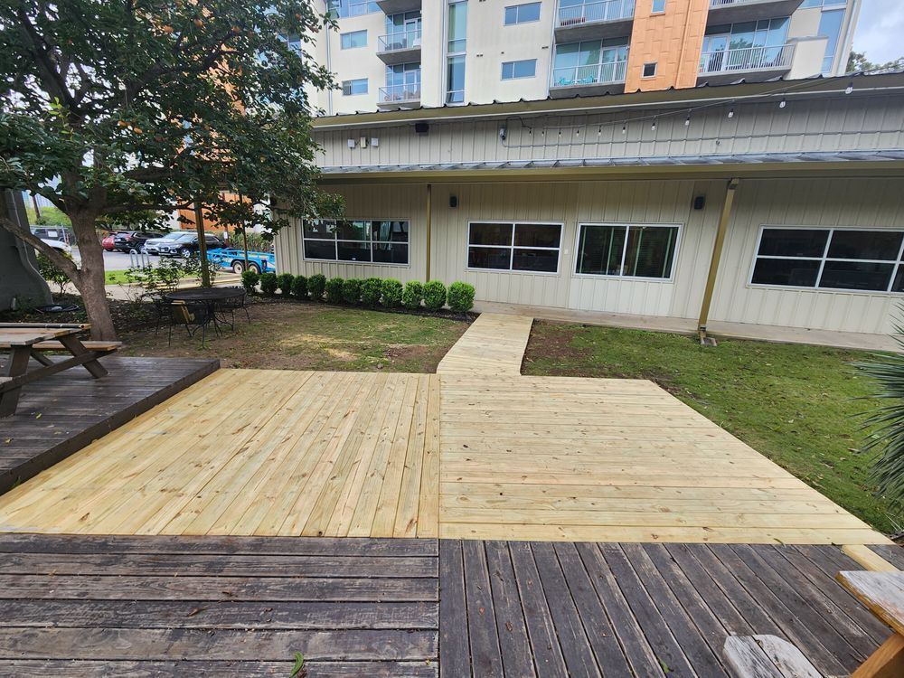 Wooden deck with new light wood and older dark wood sections, leading to a building with windows, and landscaping in an outdoor setting.