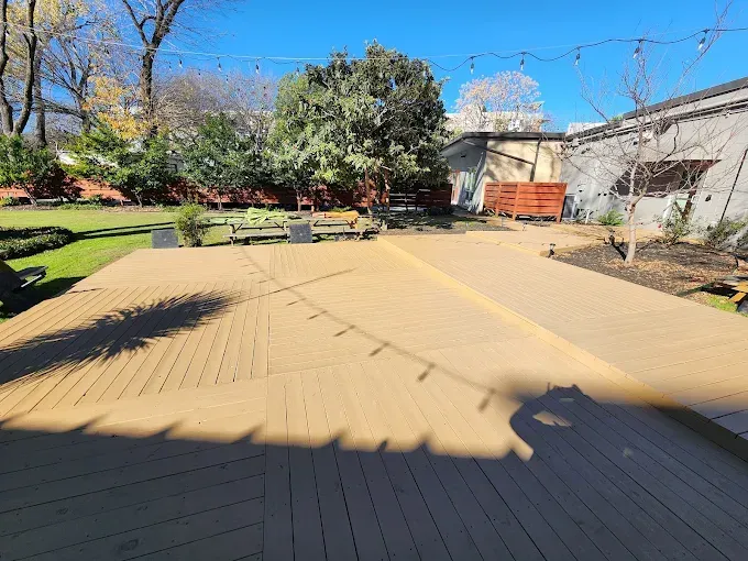 A large wooden deck in a backyard. Strings of lights hang above. A grassy area and trees are in the background.