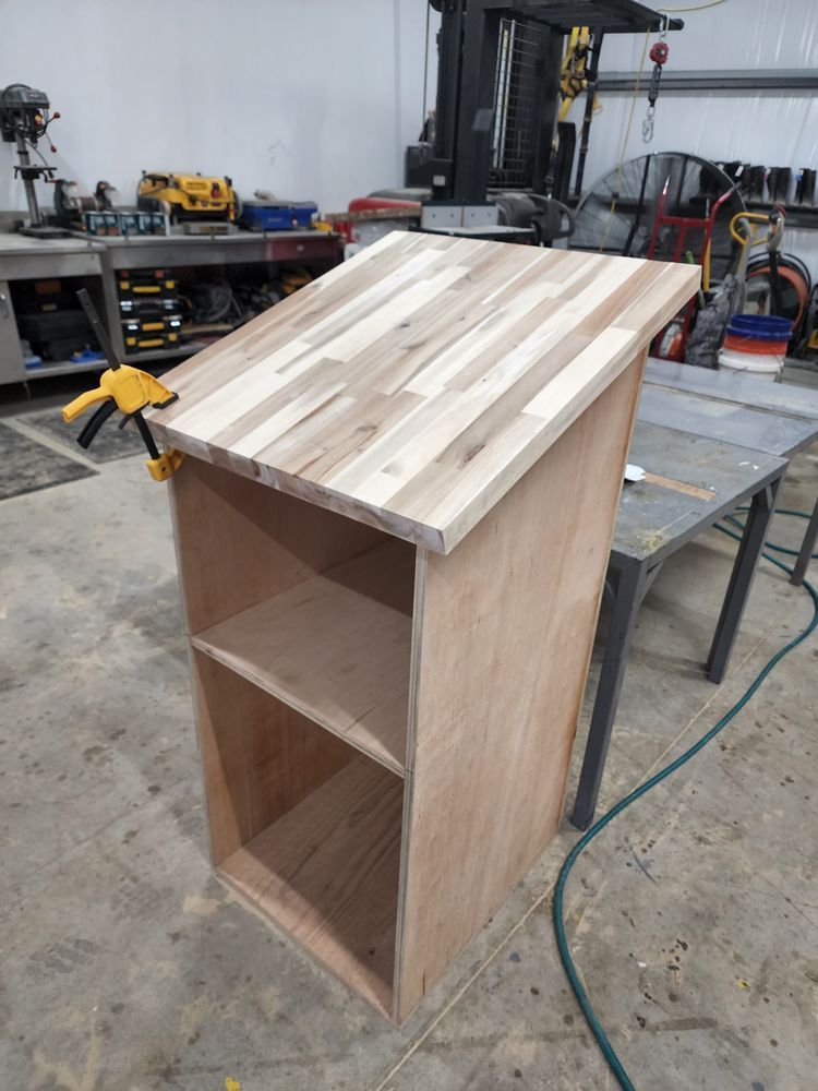 A wooden lectern under construction in a workshop. The top is made of light wood planks, and a clamp holds it in place.