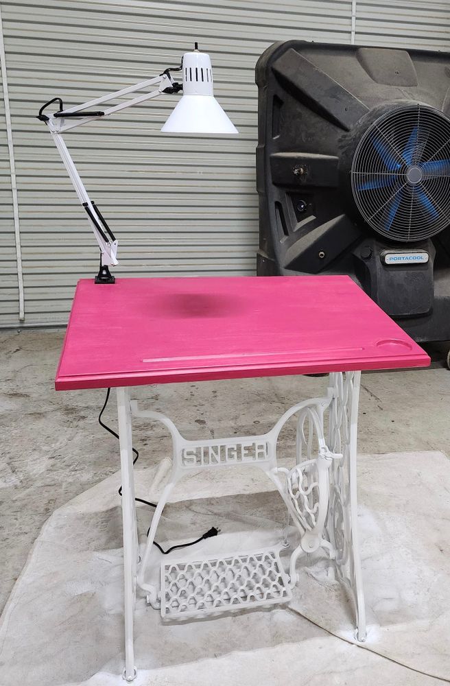 A pink sewing table with white Singer base, adjustable lamp, and a large black fan in the background.