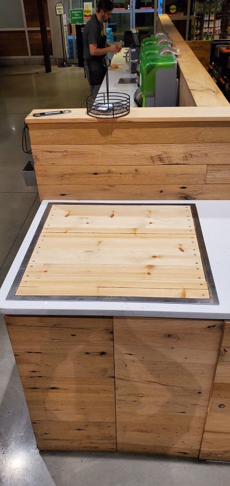 Wooden counter with a recessed wooden surface and stainless steel trim. A person works behind the counter with green containers.