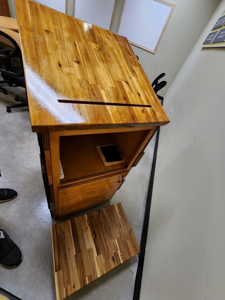 Wooden lectern with a sloped top, a slot for papers, and a missing lower panel, in a room.