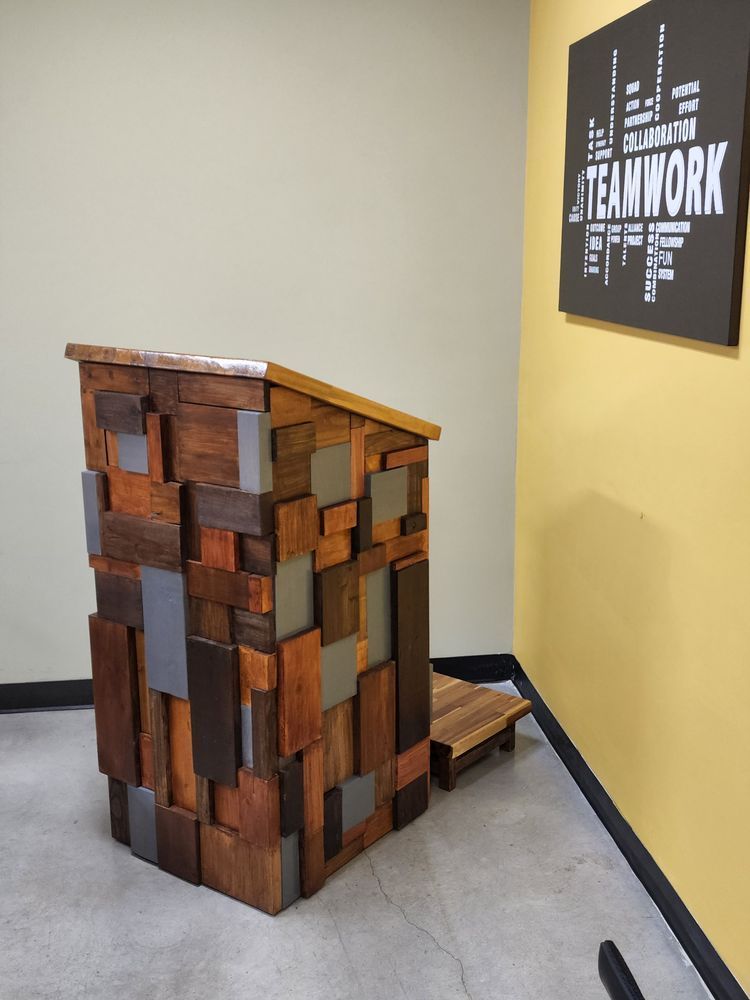 A wooden lectern made of various wood pieces with a step-stool. Teamwork sign hangs on a yellow wall in the background.
