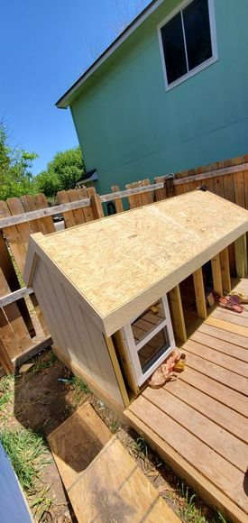 A partially constructed dog house on a wooden deck in a backyard. The house is made of light-colored wood with an open doorway.